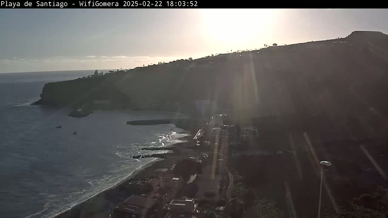 A high-angle, sunlit view shows a dark-colored beach, a calm ocean, and a cliffside town built into a hillside, under a mostly clear sky.