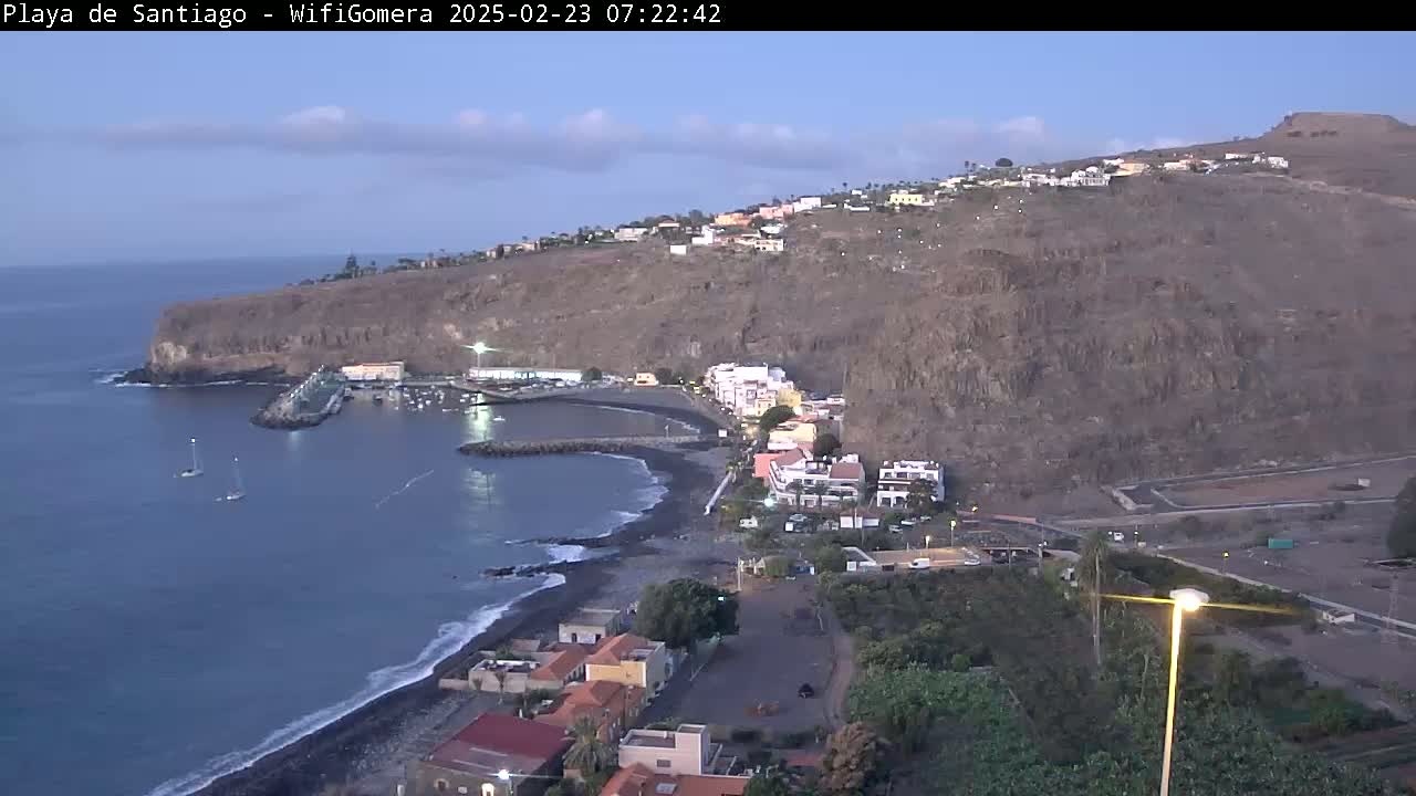 A coastal town nestled against a steep, rocky hillside overlooks a calm bay with a few sailboats at dusk.