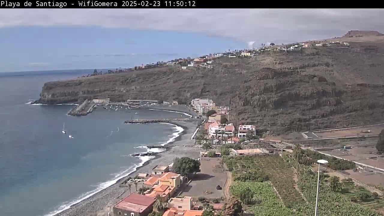 A partly cloudy day overlooking a dark-sand beach and a small town nestled against a steep, rocky hillside next to a calm ocean.