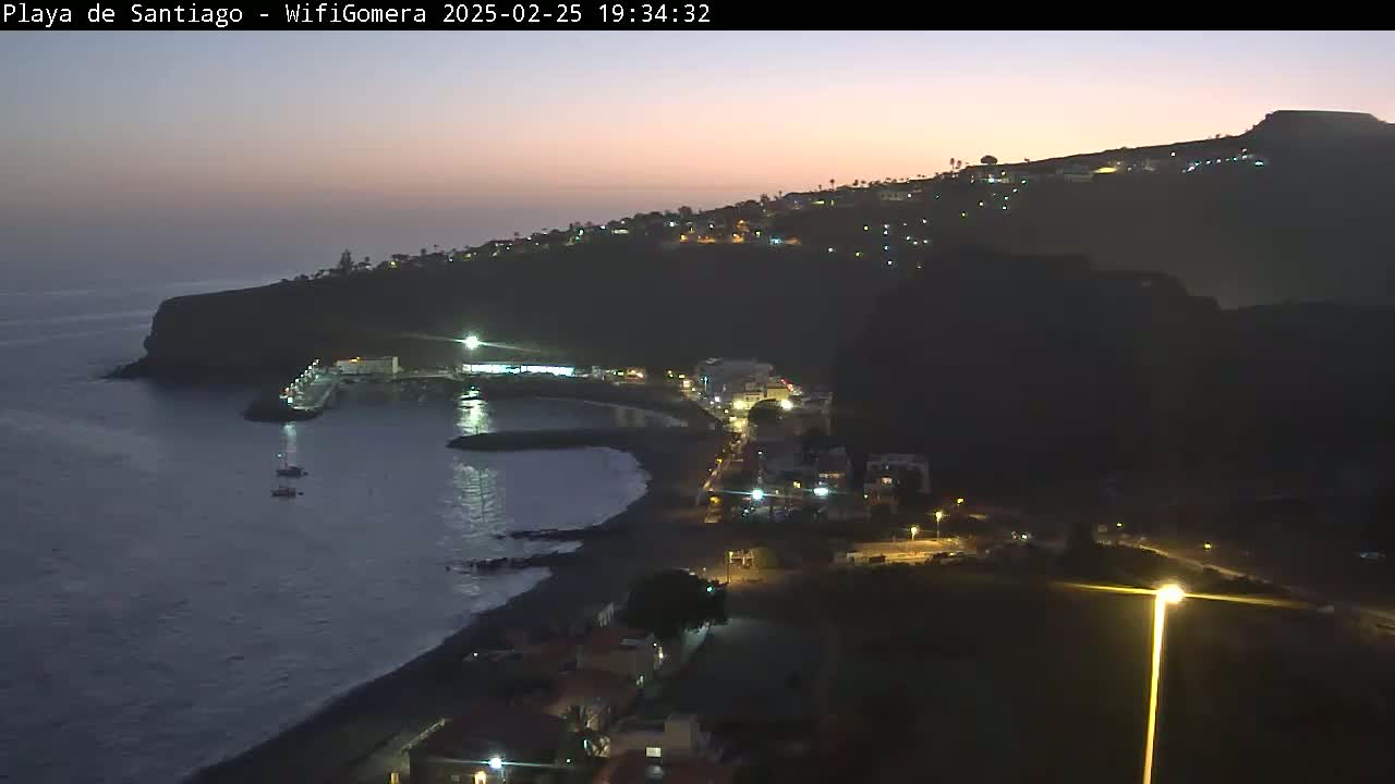 A nighttime coastal scene shows a dark, rocky cliff with a small town nestled along its edge, illuminated by streetlights, overlooking a calm, dark ocean with a few small boats.