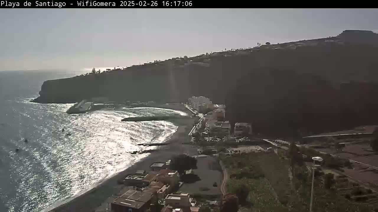 A black sand beach is partially visible below a dark cliffside town under a mostly clear sky, with somewhat choppy water lapping at the shore.