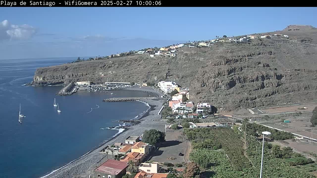 A coastal town nestled against a rocky hillside overlooks a calm bay with a few sailboats under a clear blue sky.