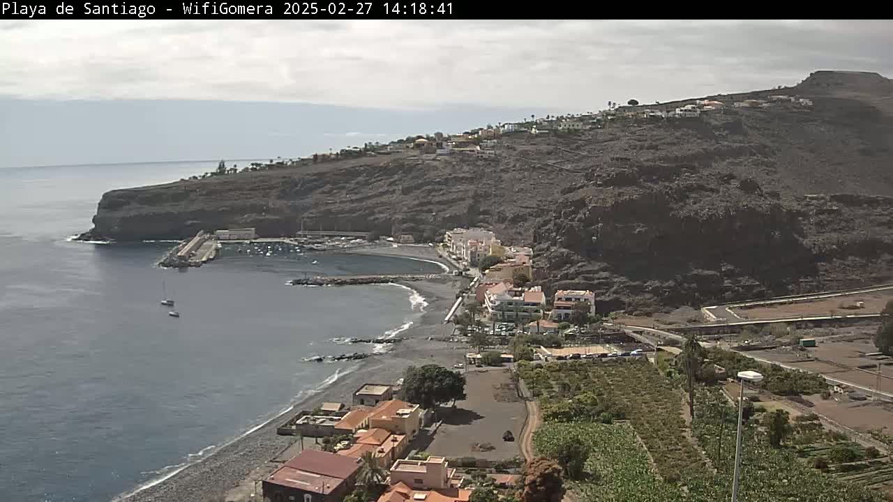 A coastal town nestled against dark volcanic hills overlooks a calm bay with a few sailboats, under a mostly cloudy sky.