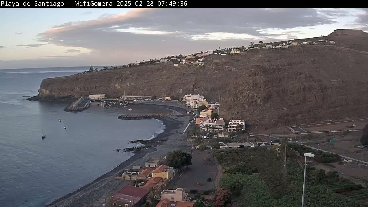 A coastal town nestled against a rocky hillside, overlooking a dark-sand beach and calm ocean under an overcast sky.