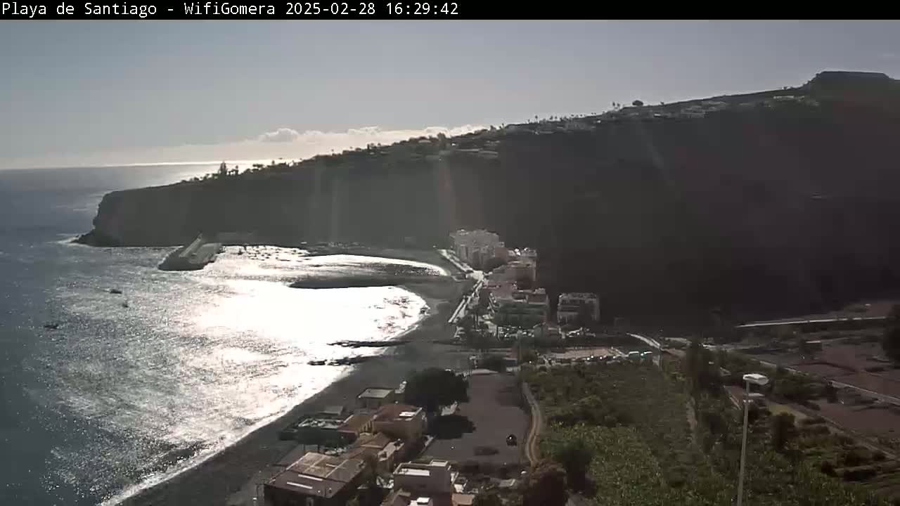 A partly sunny day shows a dark volcanic coastline with a small town and black sand beach next to a calm ocean.