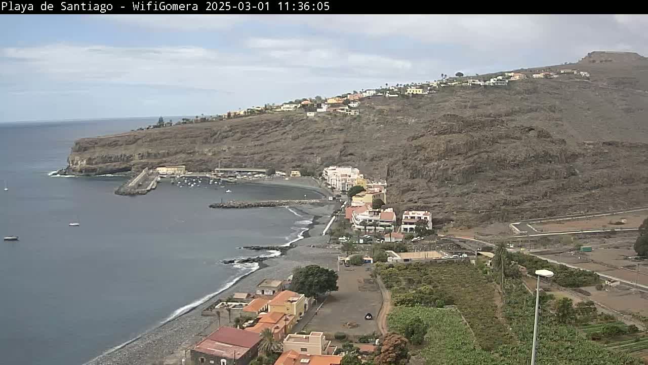 An overcast day reveals a coastal town nestled against a dark, rocky hillside, with a small, dark-sand beach and a calm bay containing several small boats.
