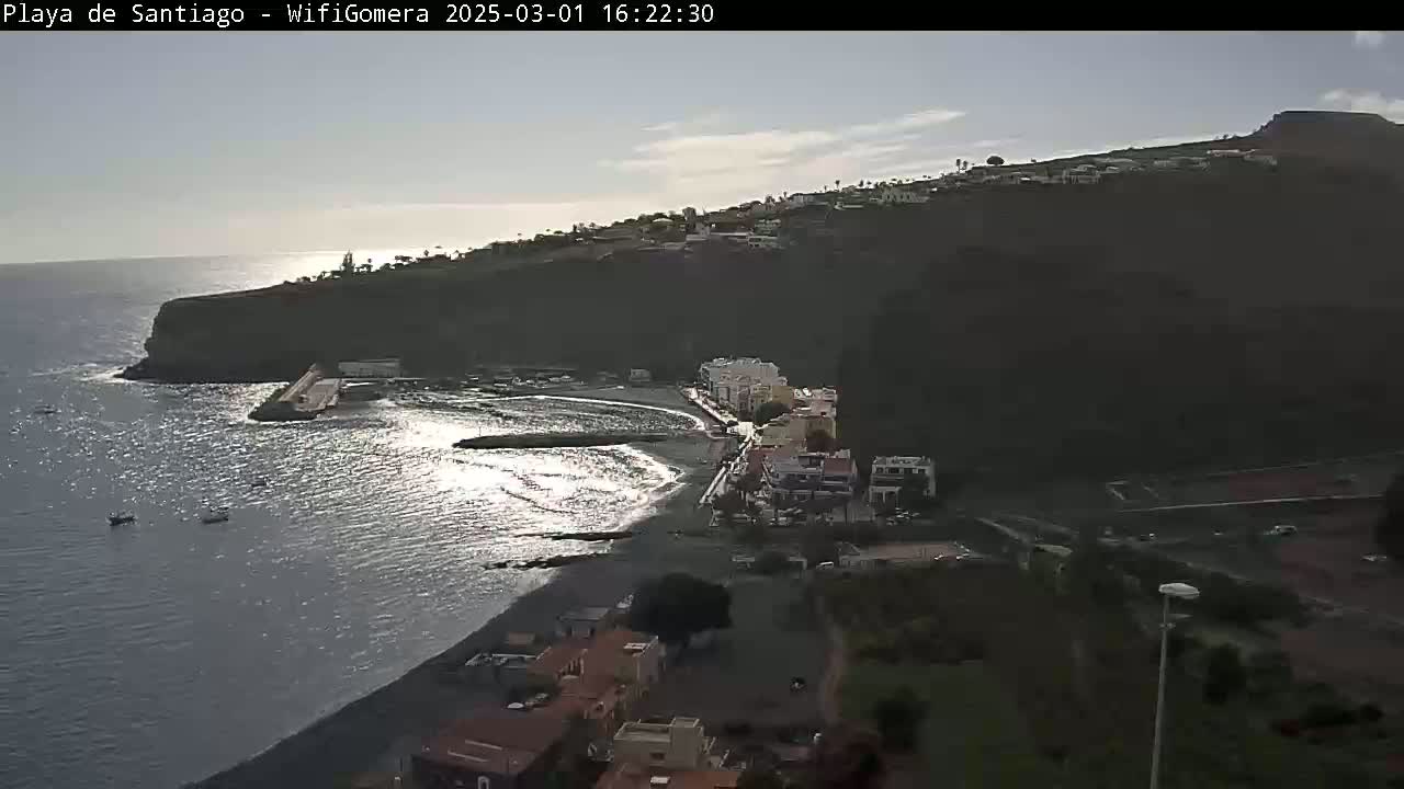 A mostly sunny day overlooking a dark-sand beach and small coastal town nestled against a steep cliffside, with several small boats visible in the calm ocean.