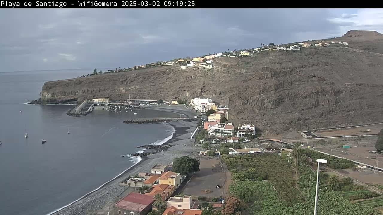 A coastal town nestled against a dark, rocky hillside overlooks a calm bay with several small boats, under a mostly cloudy sky.
