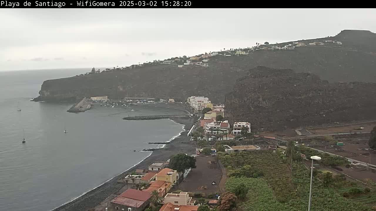 A coastal town nestled against a dark, volcanic hillside is seen on an overcast day, with a calm body of water and a few boats visible in the bay.