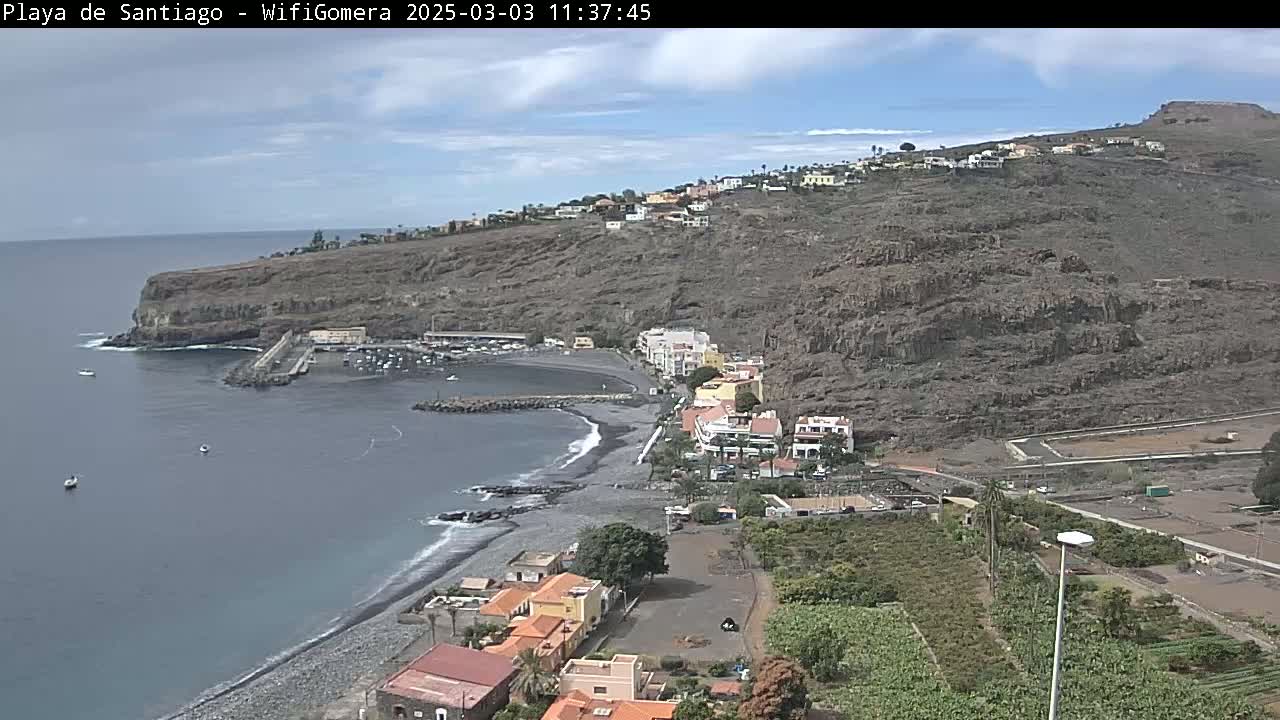A mostly sunny day reveals a dark-sand beach curving into a small harbor nestled between dark volcanic cliffs, with a small town perched above on the hillside.