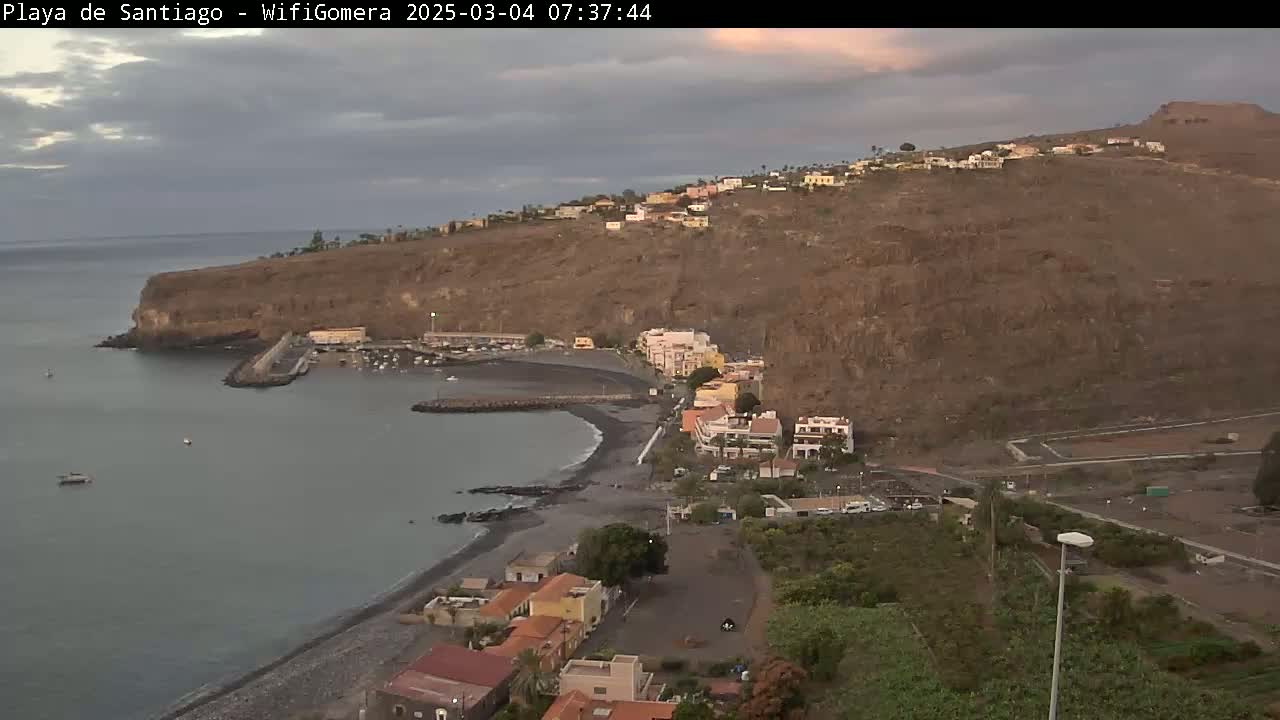 A coastal town nestled against a steep, brown hillside overlooks a calm bay with a small harbor under an overcast sky.