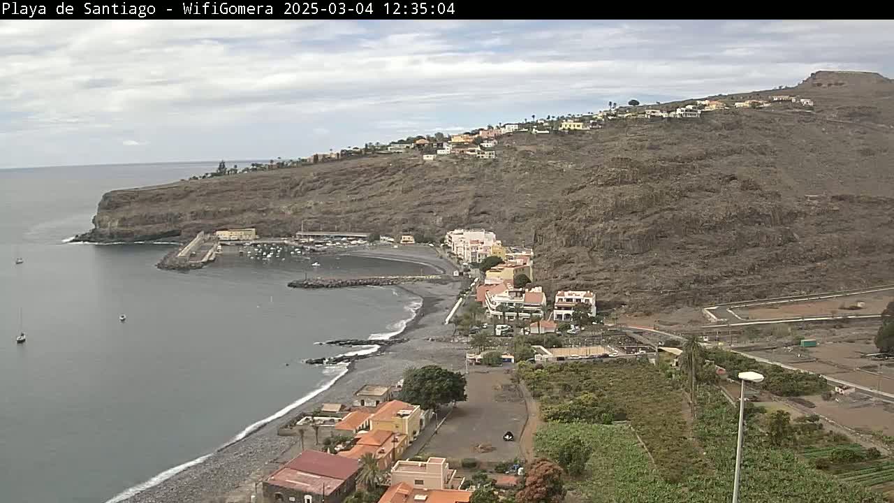 A dark-gray, pebble beach curves around a small harbor nestled beside a village built into a rocky hillside under a mostly cloudy sky.