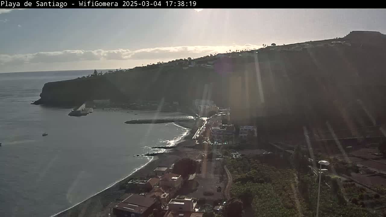 A coastal town nestled against a dark, rocky hillside is bathed in bright sunlight, with a calm sea stretching out to the horizon.