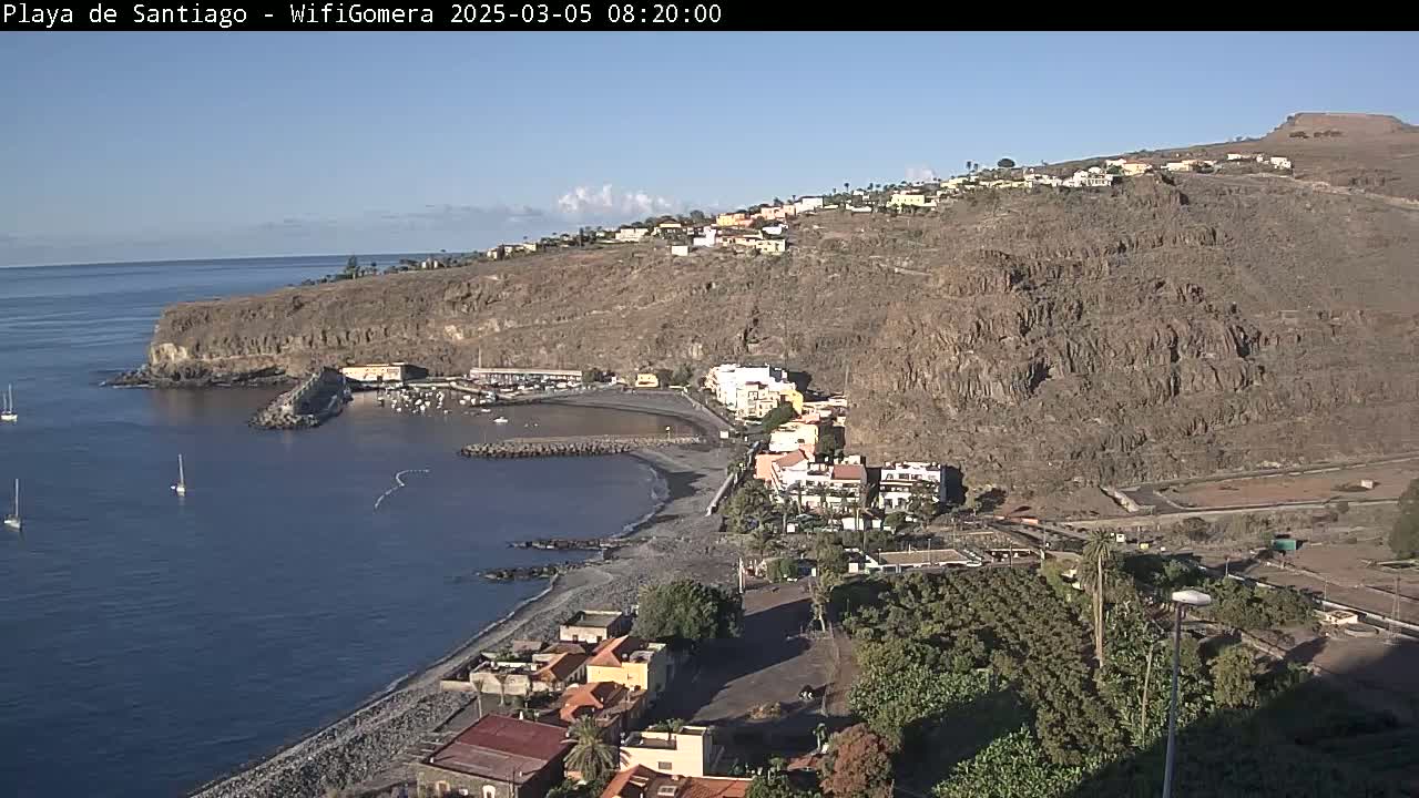 A coastal town nestled against a steep, rocky hillside, overlooking a calm bay with several sailboats, under a clear, sunny sky.