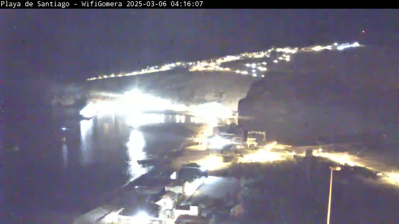A nighttime, high-angle view shows a dark body of water reflecting lights from a town situated on a hillside, with a road visible leading away from the water's edge.