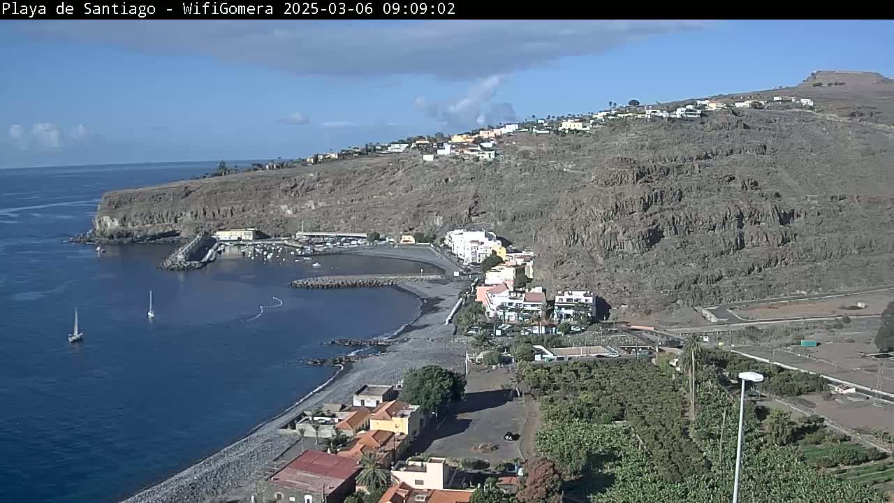 A coastal town with a small harbor nestled against a steep, rocky hillside under a mostly sunny sky.