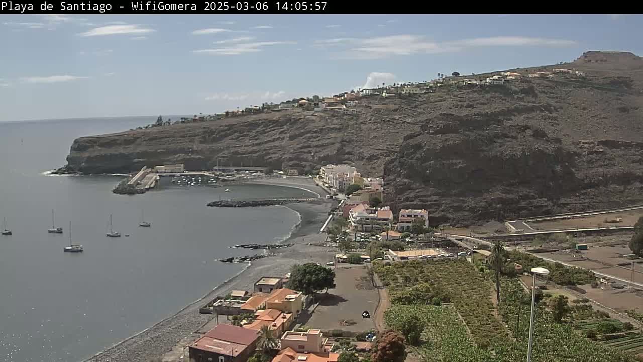 A mostly sunny day shows a dark-sand beach curving into a small bay with several sailboats at anchor, backed by a village nestled against a rocky, dark-brown hillside.