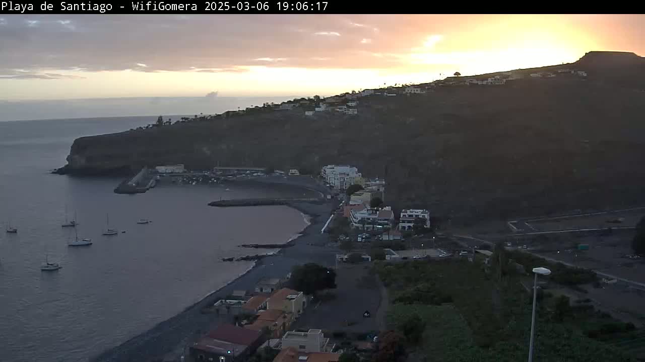A coastal town nestled against a dark volcanic hillside at sunset, with several sailboats in a calm bay.