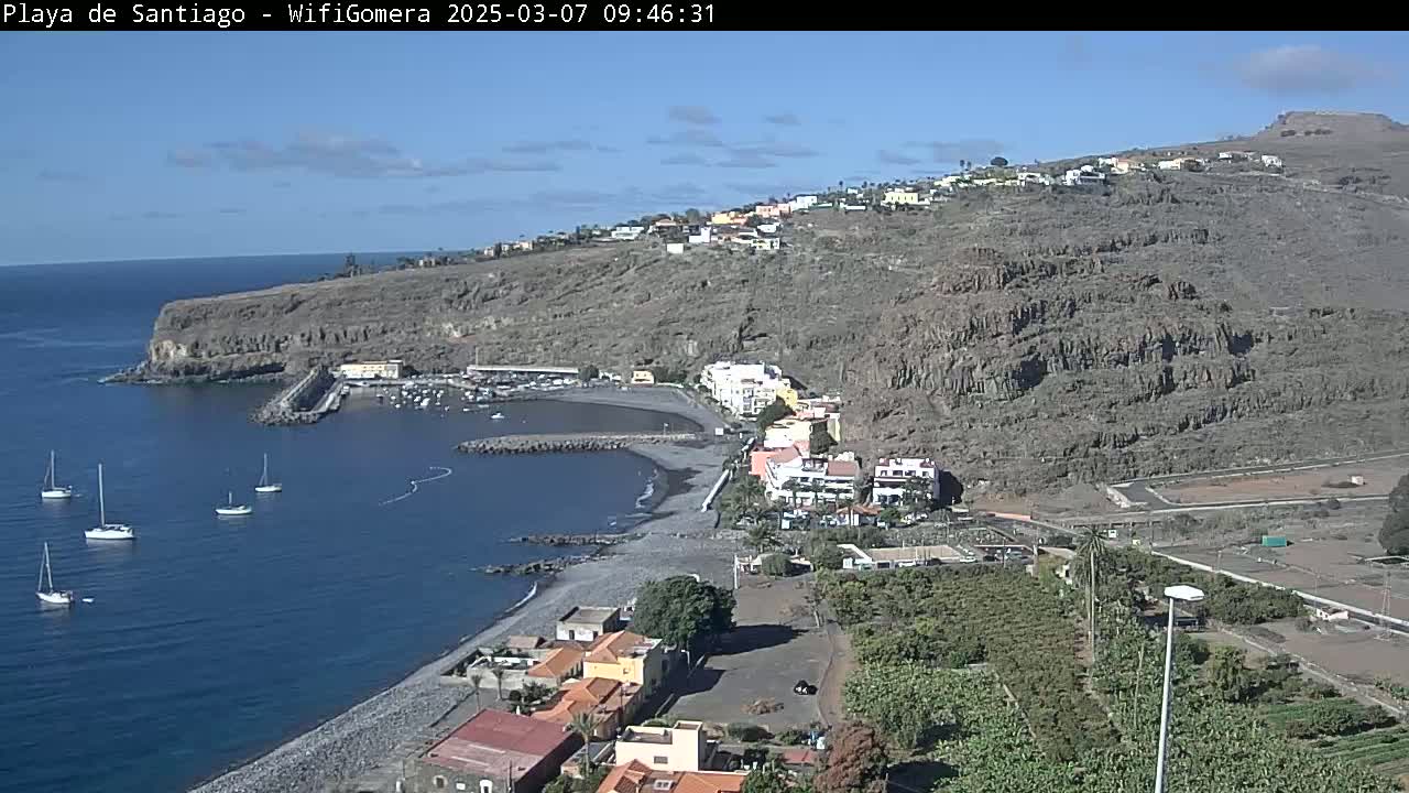 A coastal town with a small harbor and a dark-sand beach is nestled against a steep, rocky hillside under a clear blue sky.