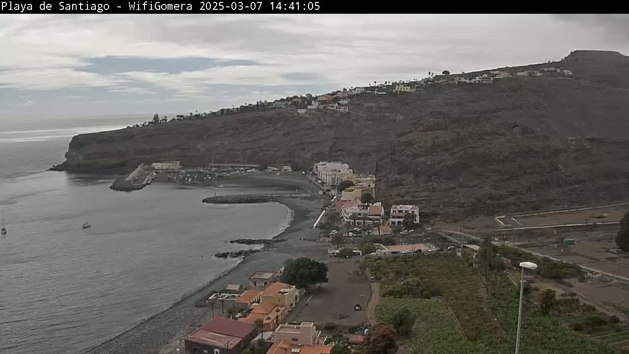 A coastal town nestled against a dark, rocky hillside on a cloudy day, with a calm ocean and a few boats visible.