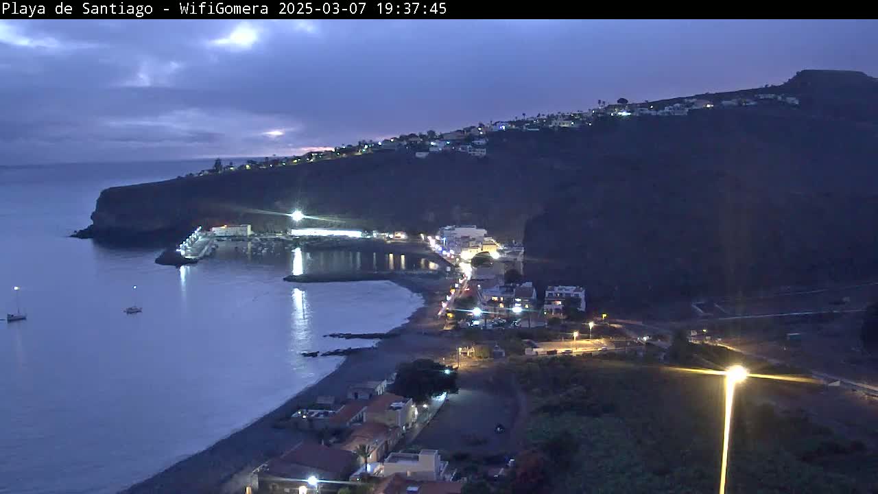 A coastal town at twilight is nestled against a dark cliffside, with a small harbor and a few boats visible in the calm water.