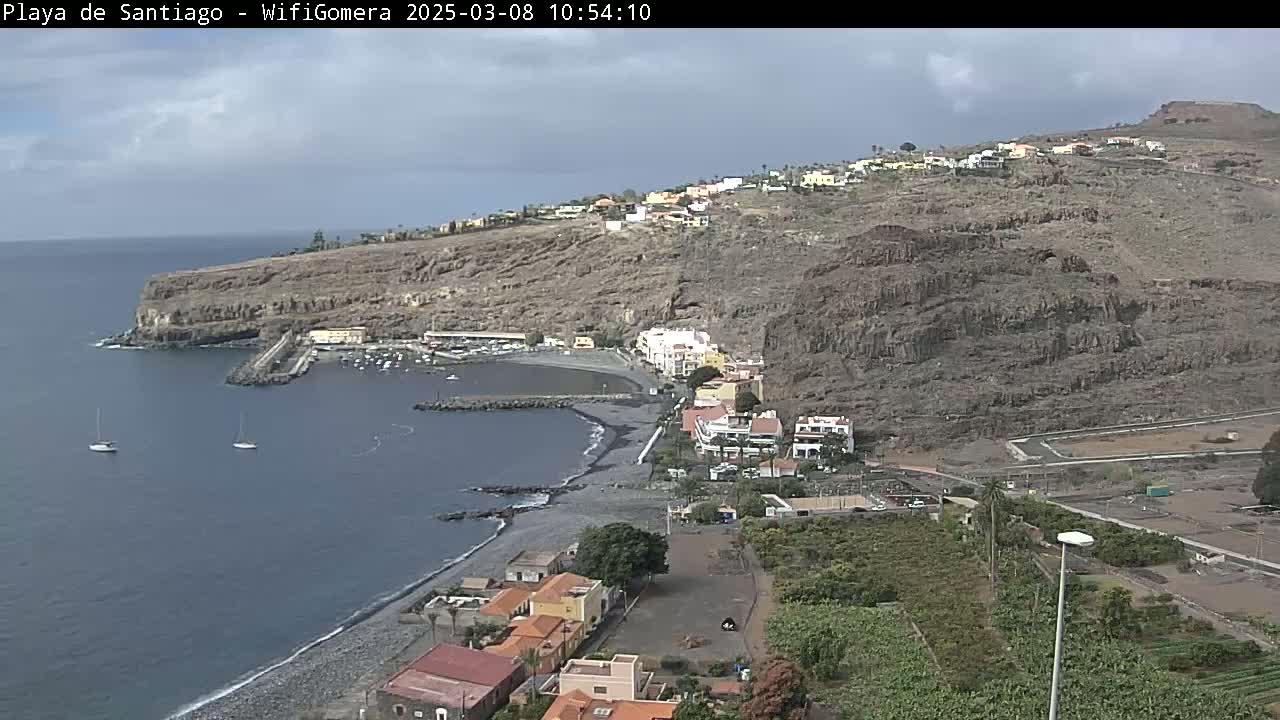 A coastal town nestled on a dark, rocky hillside overlooks a calm bay with a few sailboats under a partly cloudy sky.