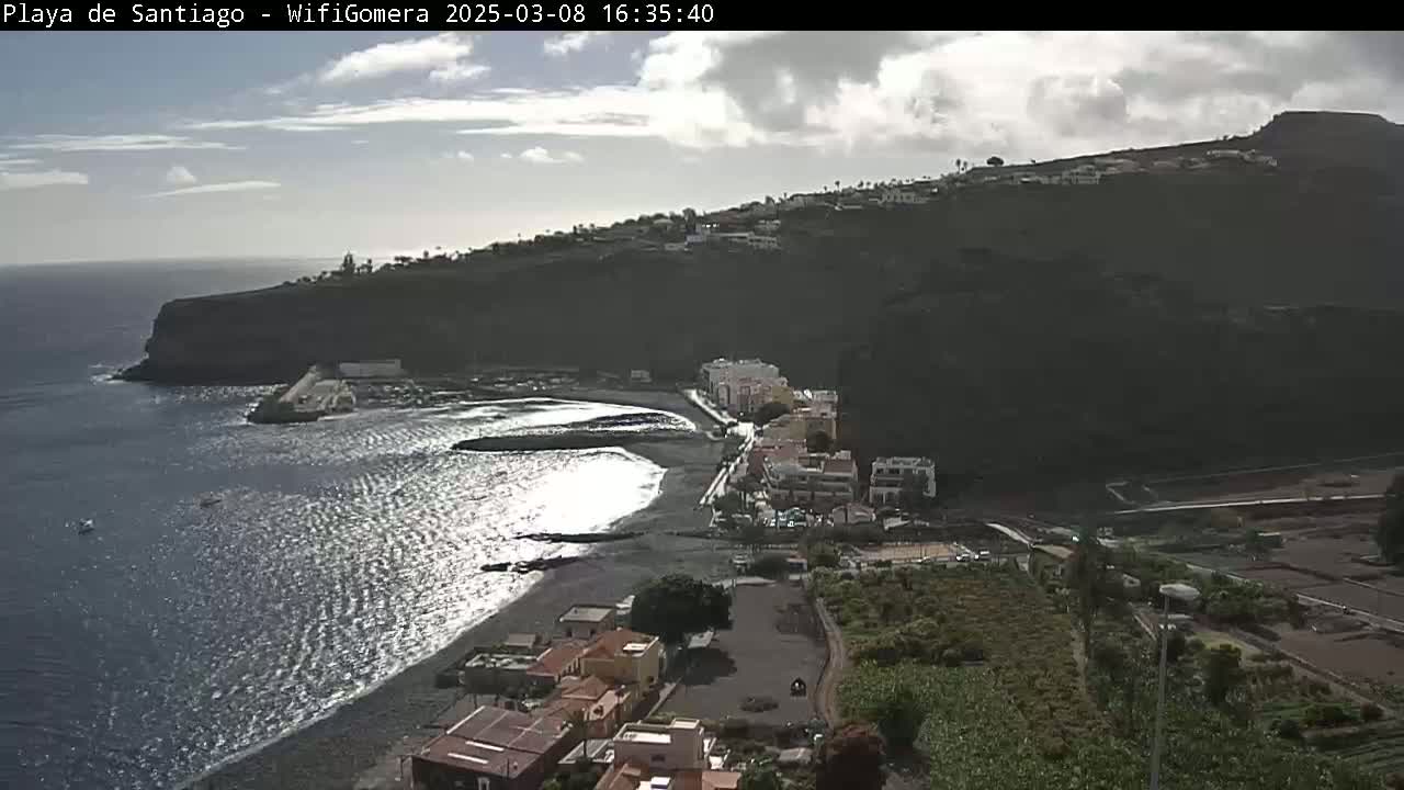 A partly cloudy day reveals a coastal town nestled against a dark cliffside, with a dark-sand beach lapping against the ocean.