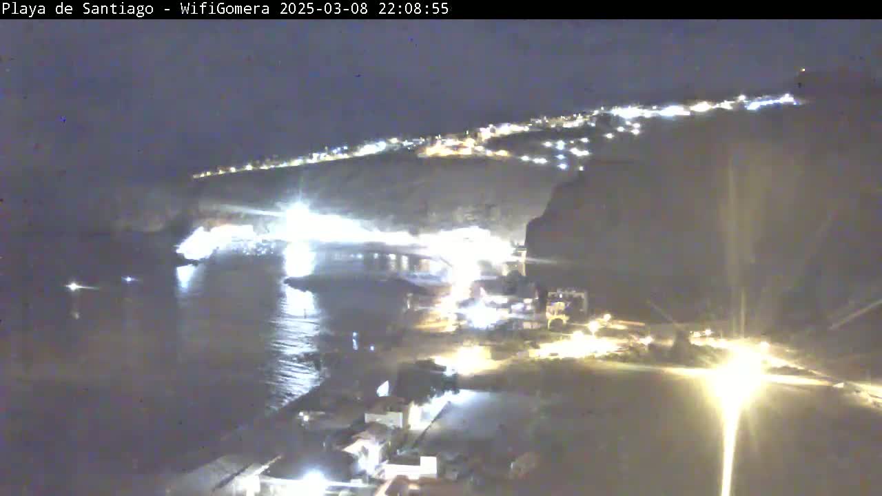 A nighttime coastal scene shows a dark body of water reflecting lights from a town built into a hillside.