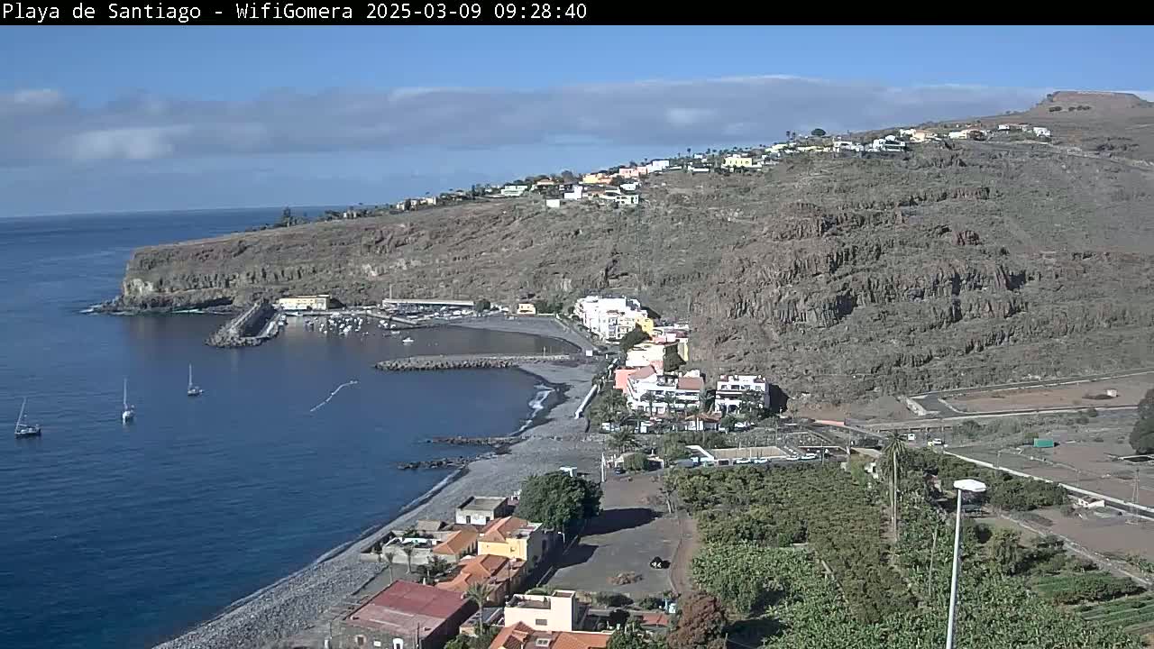 A mostly sunny day overlooks a dark-sand beach and small harbor nestled between a rocky, volcanic hillside and calm ocean water with a few sailboats.