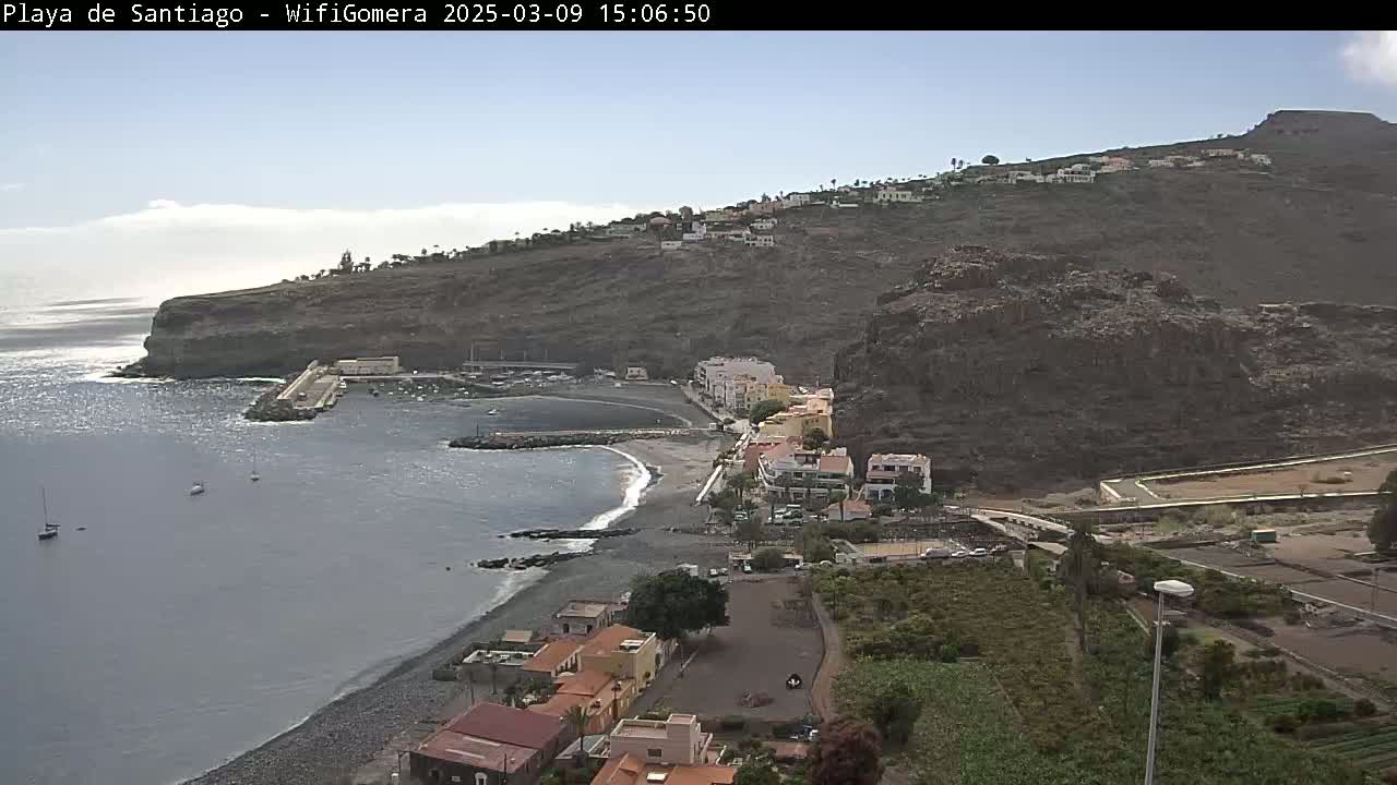 A mostly sunny day overlooking a small, dark-sand beach nestled in a cove next to a village built into a steep, rocky hillside.