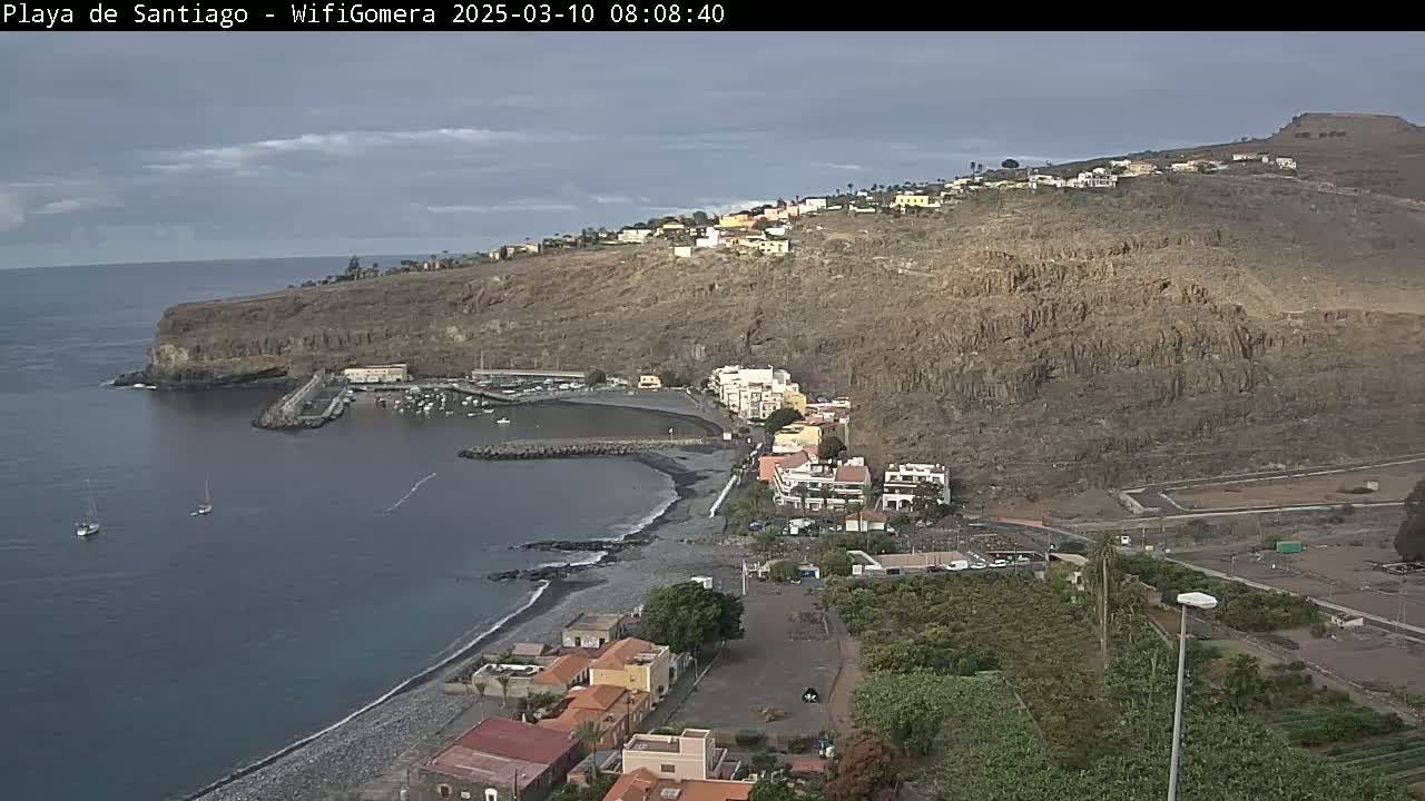 An overcast day reveals a coastal town nestled between a dark-colored beach and a steep, rocky hillside with sparse vegetation; a small harbor is visible in the bay.