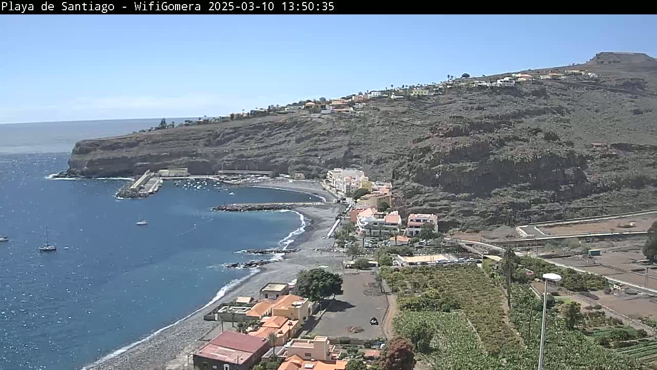 A coastal town nestled on a rocky hillside overlooks a calm, blue ocean with a small, dark-sand beach and a harbor, under a clear, sunny sky.