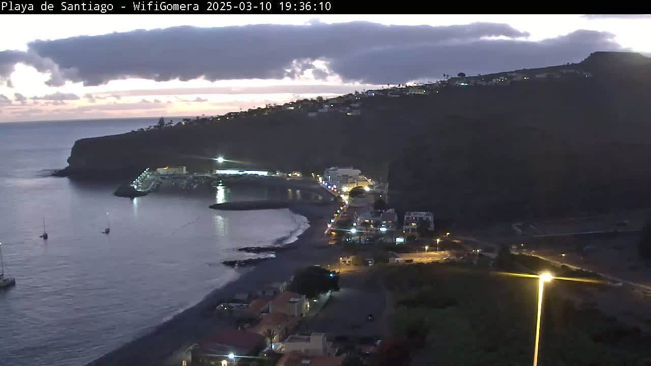 A dark, mostly cloudy evening scene shows a dark, rocky coastline with a small, dark beach, a few boats in the water, and a town with lights on a hillside overlooking the ocean.