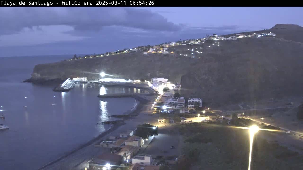 A coastal town at twilight is nestled against a dark hillside, with a calm, dark-blue ocean reflecting lights from the buildings and a small harbor.