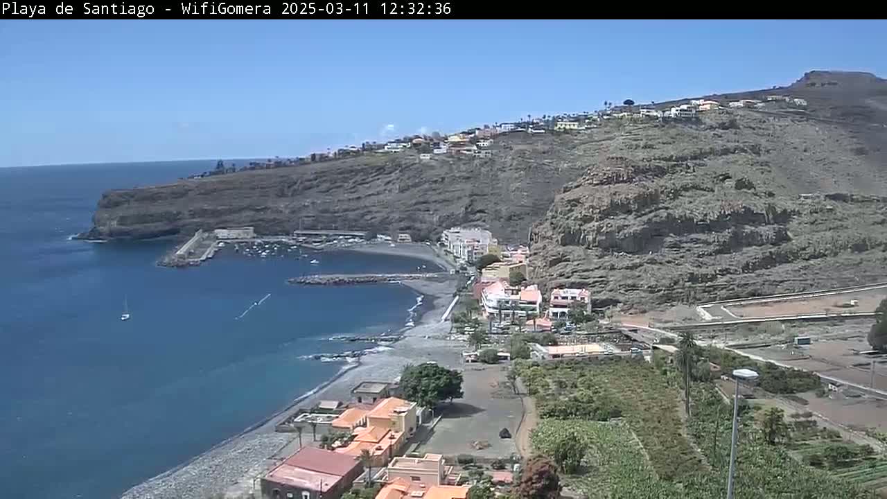 A coastal town nestled against a dark, rocky cliffside overlooks a calm, blue ocean with a small harbor and a dark-sand beach under a clear, sunny sky.