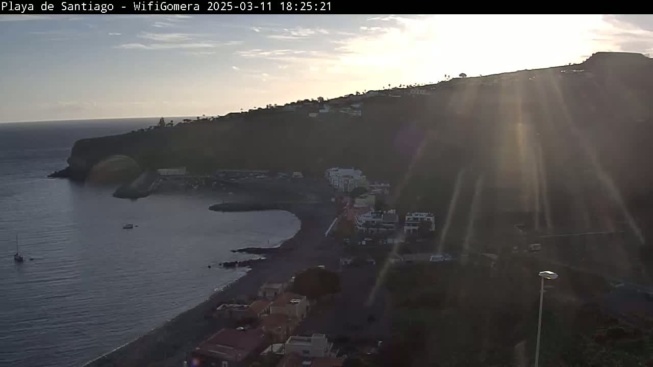 A coastal town nestled on a dark hillside overlooking a calm bay at sunset, with sunlight rays streaming down the hill.