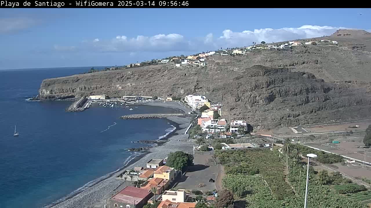 A coastal town with a dark sand beach and small harbor is nestled against a steep, rocky hillside under a mostly sunny sky.