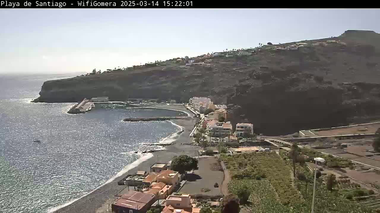 A coastal town with a small, dark-sand beach and a calm ocean under a mostly clear sky is seen from an elevated viewpoint.