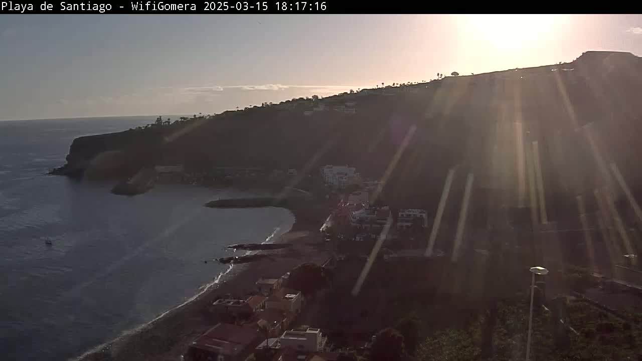 A coastal town nestled on a hillside overlooking a dark-blue ocean, bathed in the bright rays of a setting sun.