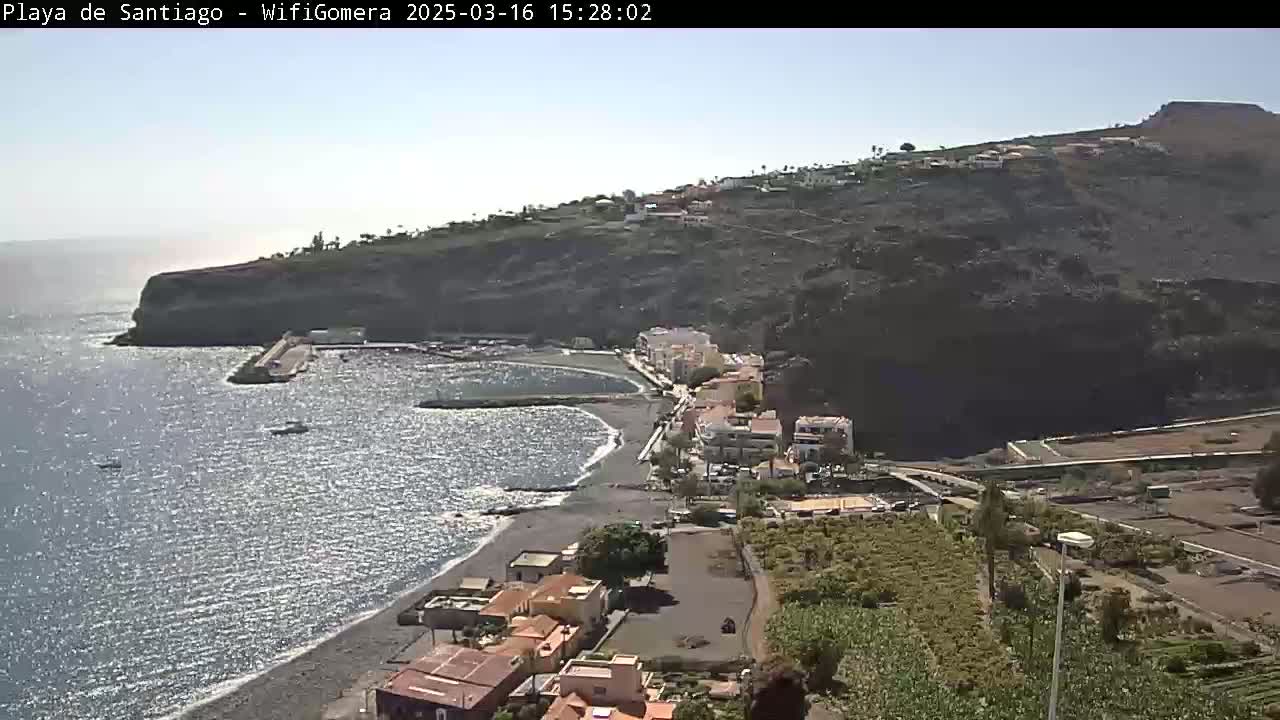 A coastal town nestled against a dark, rocky cliffside on a sunny day, with a small harbor and dark-sand beach bordering a calm ocean.