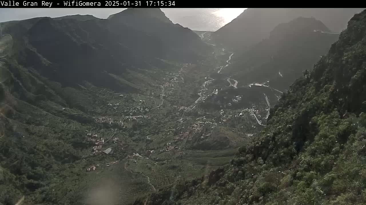 A hazy, sunlit view overlooks a valley town nestled between steep, verdant mountainsides, with a winding road visible below.