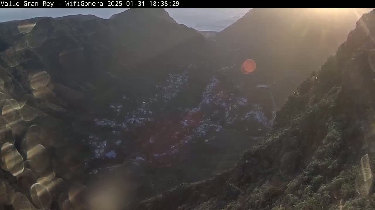 A hazy sunset illuminates a valley town nestled between steep, dark mountains.