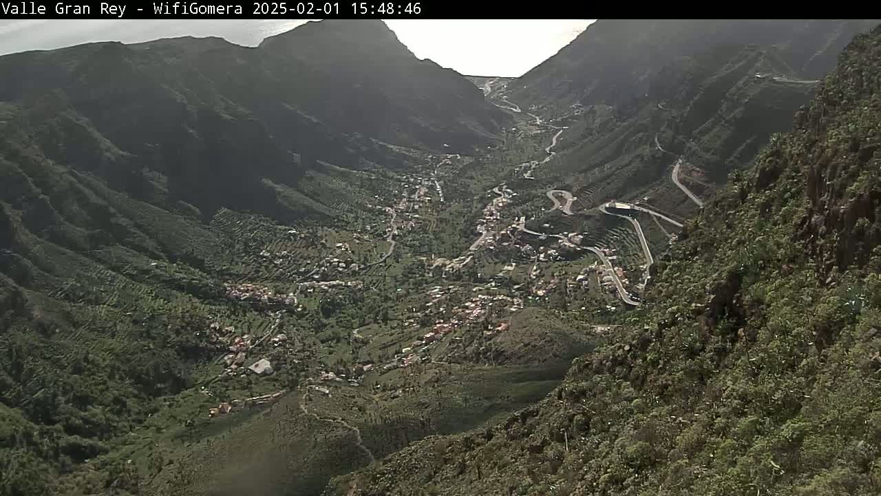 An aerial view shows a valley town nestled within a mountainous region under bright, sunny conditions.