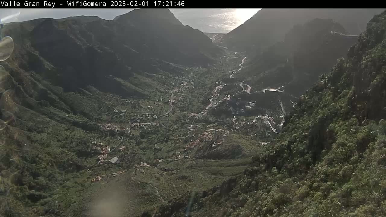 A high-angle, long shot shows a sunlit valley nestled between steep, dark green mountains, with a winding road visible through the valley.