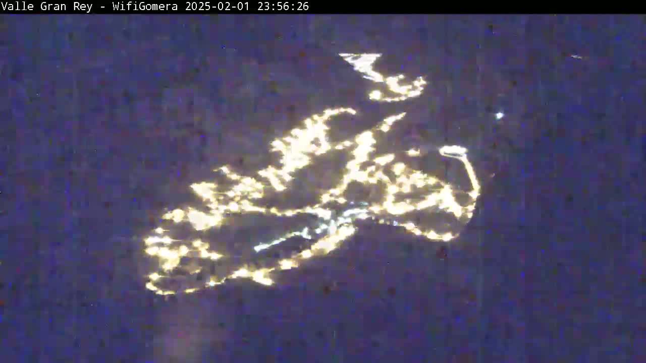 A nighttime aerial view shows a cluster of lights spread across a dark, possibly mountainous, landscape.