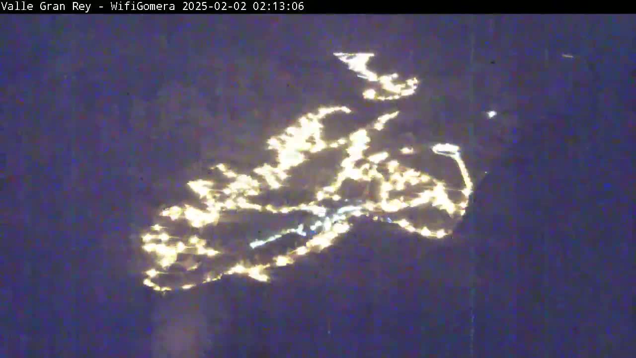 A nighttime aerial view shows the lights of a town or city reflected on a dark body of water.