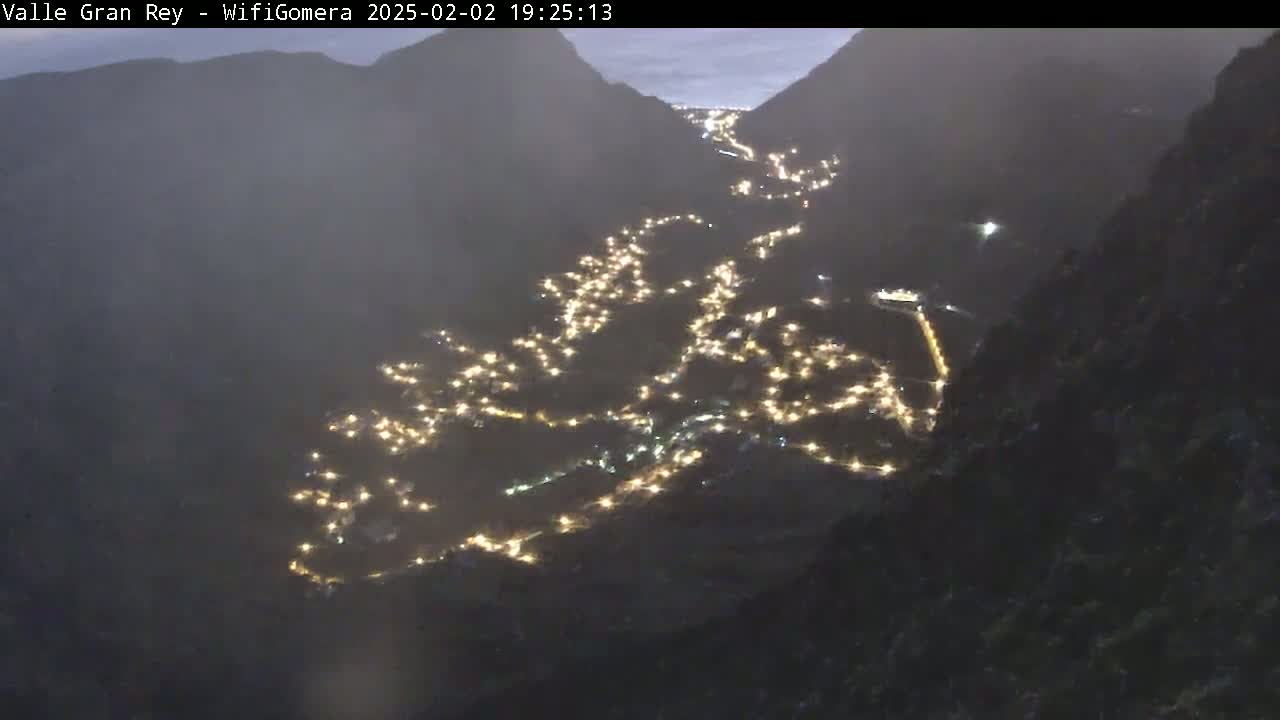 A valley town at night, nestled between dark mountains under a hazy, twilight sky, is illuminated by numerous small lights.