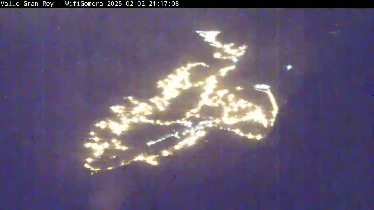A nighttime, blurry image shows a scattering of bright lights, possibly a town or city, illuminated against a dark purple sky.