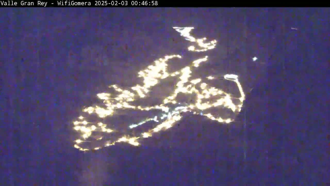 A nighttime, blurry aerial view shows the lights of a town reflected on a dark body of water.