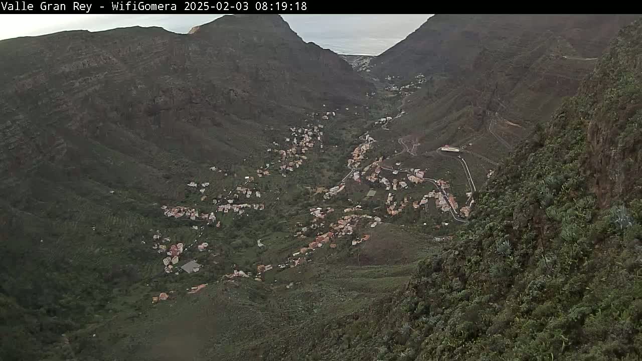 A high-angle view shows a village nestled in a verdant valley between steep, dark mountains under an overcast sky.